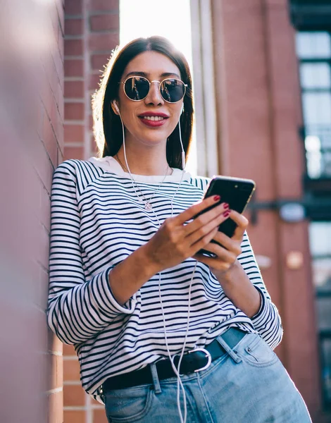 Half length portrait of millennial hipster girl enjoying music podcast while waiting friend at urban setting with modern mobile phone with radio app, positive female testing electronics headphones