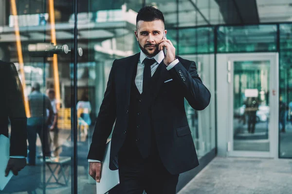 Focused bearded guy in suit looking at camera and having smartphone conversation while standing outside contemporary office building with glass walls