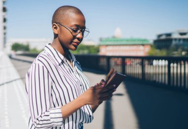 Kentteki köprü kenarında otururken akıllı telefon kullanan ve mesajları kontrol eden çizgili tişört ve kot giyen modern Afrikalı Amerikalı kadınların yan görüntüsü