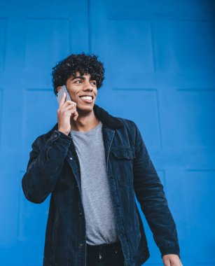 Gülümseyen, siyah ceketli ve kot pantolonlu genç, kıvırcık etnik adam caddede mavi şehrin kapısının önünde dikilirken telefonla konuşuyor.