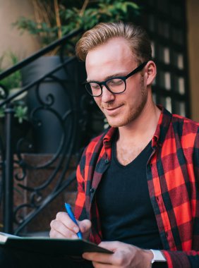 Neşeli, kızıl saçlı, gözlüklü, işaretli kırmızı ve siyah tişörtlü bir erkek. Yazın şehirde granit merdivenlerde oturan kalemle not alıyor.