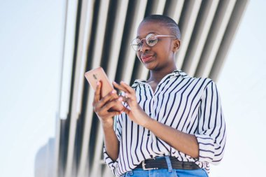 Çizgili bluz ve kot pantolonlu şık, kel, Afro-Amerikan kadın yazın köprü altında dururken akıllı telefondan mesajları kontrol ediyor.