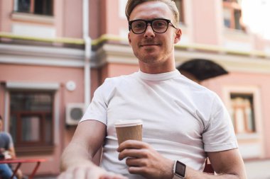 Tatmin olmuş beyaz gömlekli ve gözlüklü genç adam güneşli bir günde kafede dinlenirken bir fincan kahve içiyor.