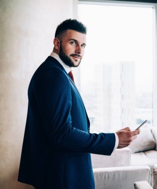 Ofisin dinlenme yerinde gülümseyen ve kameraya bakarken iş stili kıyafet taraması yapan yakışıklı yöneticinin yan görünüşü