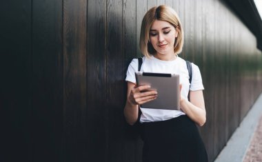 Sıradan kıyafetler giymiş genç sarışın bir kız öğrenci. Tableti karıştırıyor ve dinleniyor. Sokaklarda durup ahşap duvara yaslanıyor.