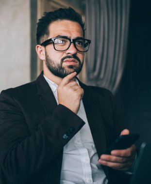 Resmi takım elbiseli ve gözlüklü düşünceli bir adam. Ortağıyla akıllı telefon mesajlaşması yapıyor. Çenesine dokunuyor.