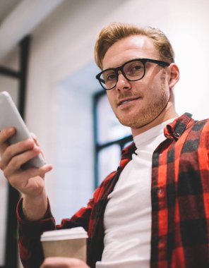 Düşünceli, sakin, stil sahibi, kahveli serbest çalışan, iş yerinde kameraya bakarken cep telefonu kullanarak haber okuyan biri.