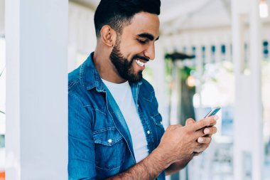 Gündüz kıyafetleri içinde gülümseyen, iyimser etnik erkek cep telefonu kullanıyor sosyal medyada arkadaşlarıyla sohbet ediyor güneşli bir günde.