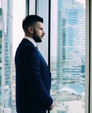 Modern şirket ofisinde çalışırken büyük pencereden dışarı bakan siyah sakallı, başarılı adamın yan görünüşü.