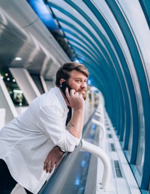 Hafif geometrik koridorda çitlere yaslanmış, cep telefonuyla konuşan hoş sakallı adamın yan görüntüsü.