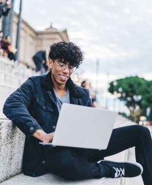 Gözlüklü mutlu etnik adam sıradan kıyafetler giyiyor. Şehir sokaklarında merdivenlerde arka planda insanlarla otururken bilgisayarını faiziyle kullanıyor.