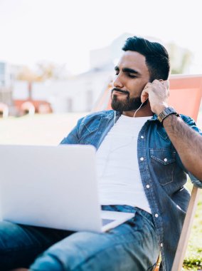Rahat rahat gülümseyen, sıradan kot gömlekli, kulaklıklı, dizüstü bilgisayarlı müzik dinleyen, güneşli bir parkta vakit geçiren genç bir erkek.