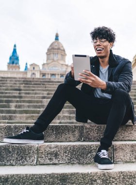Siyah ceketli, komik, modern bir erkek görüntülü sohbet için tablet kullanırken arka planda bulanık bir şekilde sokak merdivenlerinde oturuyor.