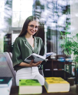 Smiling inquisitive young woman in eyeglasses leaning on armchair and holding book in modern library hall while looking at camera through glass clipart