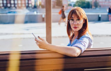 Şaşkın kızıl saçlı, sıradan giyinmiş, akıllı telefon kullanan, sosyal medyayı kontrol eden, bankta oturup kameraya bakan bir kadının arka plan görüntüsü.