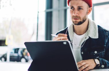 Sıcak giyimli, şapkalı, elinde gözlük ve kalemle iş yerinde bulanık arka planda yan bakan dalgın genç bir serbest erkek.