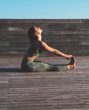 Yoga kıyafeti giymiş, bacakları esneyen ve sabah tahta çitin önünde, kereste zeminde oturan sağlıklı bir kadın.