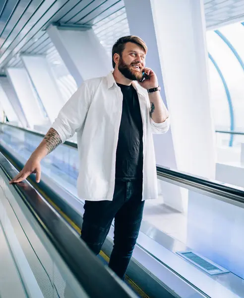 Pleasant bearded man in black t-shirt and white shirt speaking on mobile phone moving on travelator holding handrail in light geometric tunnel