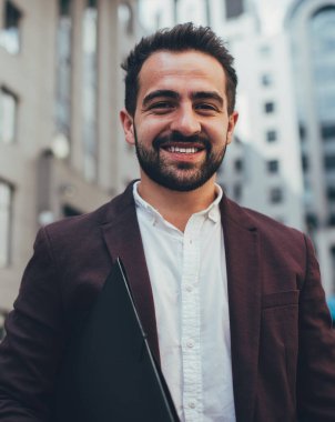 Moda kıyafetli neşeli beyaz adamın yarım boy portresi şehir planlama arka planında duruyor, genç iş adamı elinde kağıt klasör tutuyor ve başarılı kariyerinden memnun kameraya bakıyor.
