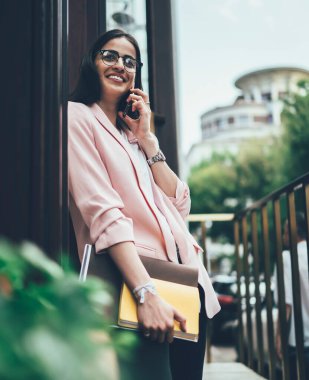 Kağıt klasörü ve günlüğü olan neşeli bir bayandan telefon görüşmesi yapan ve yaz günü binanın kapısında durup gülümseyen.