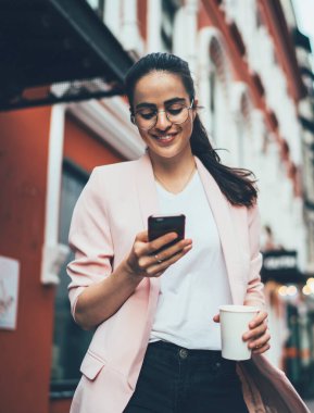 Gözlüklü, siyah saçlı, gözlüklü, şık elbiseli, beyaz perdeye konsantre olan ve cadde boyunca bulanık cephelere karşı yürürken akıllı telefonla etkileşim kuran bir kadın.