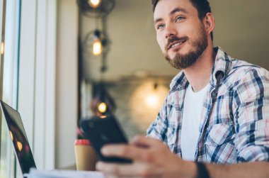 İş yerinde cep telefonuyla sörf yaparken kareli tişörtlü, güler yüzlü, düşünceli, düşünceli bir erkeğin bakışlarının altından.