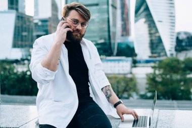 Gündelik giysiler ve yuvarlak gözlükler içinde odaklanmış bir erkek akıllı telefondan iş arkadaşıyla konuşuyor. Proje üzerinde çalışıyor. Modern şehrin sokaklarında dizüstü bilgisayarla kameraya bakıyor.