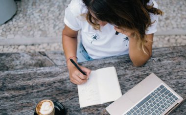 Günlük giysiler içinde bir kadın bir fincan kahve ve dizüstü bilgisayarla masada oturuyor ve telefonda konuşan boş defterlere bazı notlar yazıyor.