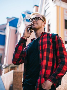 Kırmızı sakallı, gözlüklü, kırmızı ve siyah tişörtlü bir erkek yazın şehirde cep telefonundan arama yapıyor.