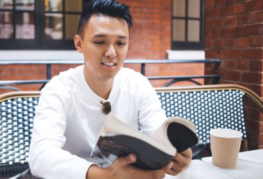 Beyaz tişörtlü yakışıklı Asyalı bir erkek kahve fincanı ile masada oturuyor ve kafede vakit geçirirken yeni bir kitap çeviriyor.