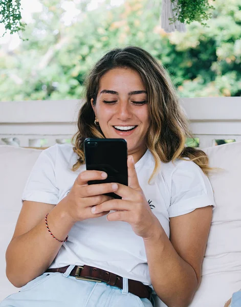 Happy young female using mobile phone and laughing excited with news from social media while sitting on sofa with pillows on terrace