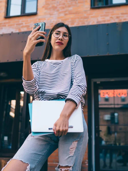 Latino student with netbook posing at urban setting for making web selfie content via front smartphone camera, millennial freelancer posing for creating media publication for sharing to social site