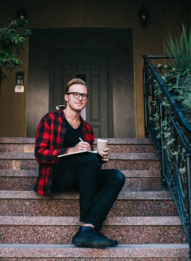 Yaratıcı gülümseyen kızıl saçlı adam kırmızı ve siyah gömlekli işaretli not defterine not alıyor ve yazın granit merdivenlerde kahveyle birlikte oturuyor.