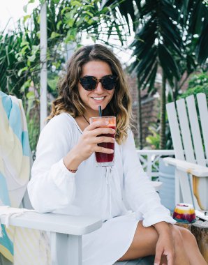 Keyifli, rahatlatıcı, güneş gözlüklü, sandalyede dinlenen ve tatil sırasında kahvaltının tadını çıkaran rahatlamış, dinlendirici bayan.