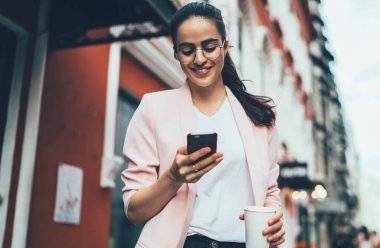 Gözlüklü, siyah saçlı, gözlüklü, şık elbiseli, beyaz perdeye konsantre olan ve cadde boyunca bulanık cephelere karşı yürürken akıllı telefonla etkileşim kuran bir kadın.