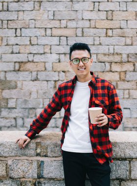 Siyah saçlı, sıradan bir erkek, gözlüklü, plastik bardaklı, gülümseyen ve kameraya bakarken eski duvarlara yaslanan.