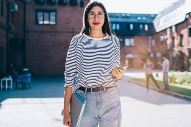 Moda kuşağı için akıllı telefonlu modaya uygun giyinmiş bayan blog yazarının yarım boy portresi boş zamanlarında üniversite kampüsünde yürüyor, açık havada eğitim için ekipmanları olan çekici bir kadın.