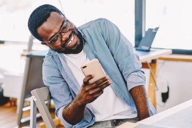 Gözlüklü neşeli siyah adam akıllı telefon ekranına bakıyor ve gülerek ofisteki masada oturuyor.