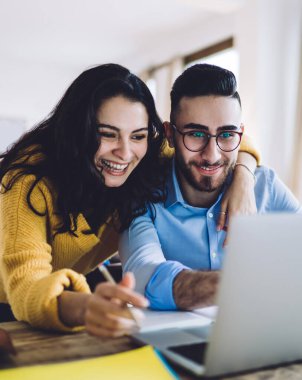 Neşeli siyah saçlı kadın süslü gözlüklü yakışıklı adama sarılıyor, işten sonra laptopta komik videolar izliyor ve evde gülüyor.