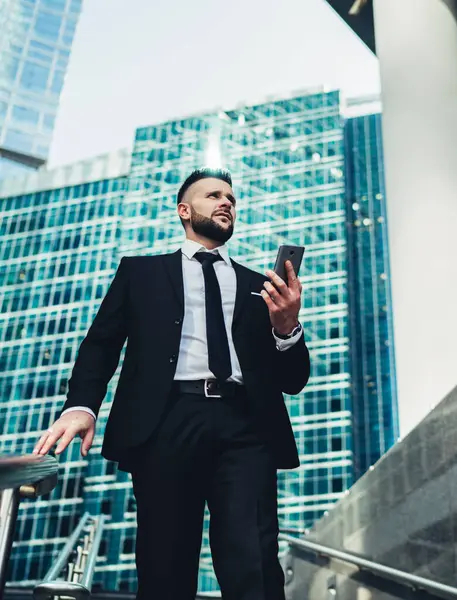 From below of bearded young executive man in classy formal black suit holding mobile phone walking down stairs in modern city with skyscrapers