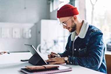 Yandan bakan genç erkek serbest grafik tasarımcı, kalın kıyafetler giyiyor, gözlük takıyor ve parlak beyaz odada tablet üzerinde çalışıyor.