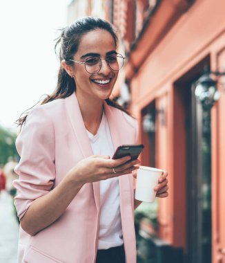 Düşük açılı, gözlüklü, sıcak içecekli zarif bir kadın cadde boyunca yürürken kameraya bakıp gülecek ve bulanık dış görünüşe karşı akıllı telefonuyla etkileşime girecek.