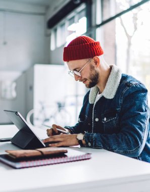 Yandan bakan genç erkek serbest grafik tasarımcı, kalın kıyafetler giyiyor, gözlük takıyor ve parlak beyaz odada tablet üzerinde çalışıyor.