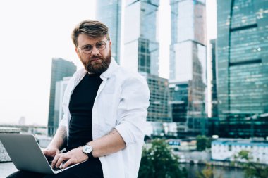 Şehirdeki iş sahasında arka planda gökdelenler ile çalışan erkek serbest yazar portresi. Netbook 'ta güvende olmak için antivirüs sistemini güncelleyen klasik gözlüklü hipster adam.