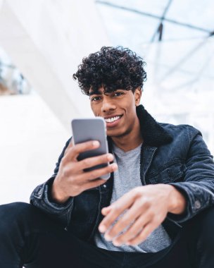 Beyaz modern binada otururken kameraya bakan, kıvırcık saçlı, şık giyinmiş, mutlu, Afro-Amerikan bir adamdan.