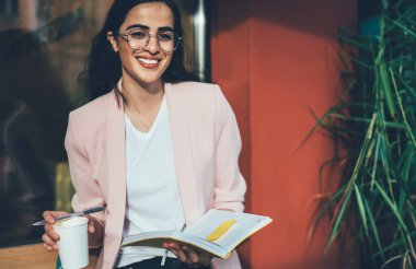 Milenyum kadını, gözlüklü ve resmi giysili, elinde hazır kahveyle ahşap bankta oturmuş notları kontrol ederken kameraya gülümsüyor.