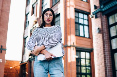 Şehir merkezinde teknoloji teçhizatıyla poz veren çekici kız öğrencinin yarı boy portresi. Arka planda şehir binaları olan güzel bir kadın, dışarıda dizüstü bilgisayar tutan gözlüklü bir kadın.