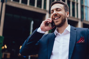 Düşük açılı mutlu lider resmi olarak gülerken gözleri kapalı caddede duruyor ve şehir merkezinde akıllı telefondan konuşuyor.