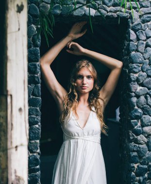 Çekici, düşünceli, romantik bir kadın. Açık renk saçlı, kameraya bakıyor. Bali 'nin taşlı kapısında ellerini kaldırmış duruyor.