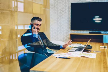 Zarif takım elbiseli başarılı bir adamın iş yerinde dizüstü bilgisayarı izlemek için akıllı telefon kullanarak partneriyle konuştuğu camdan bak
